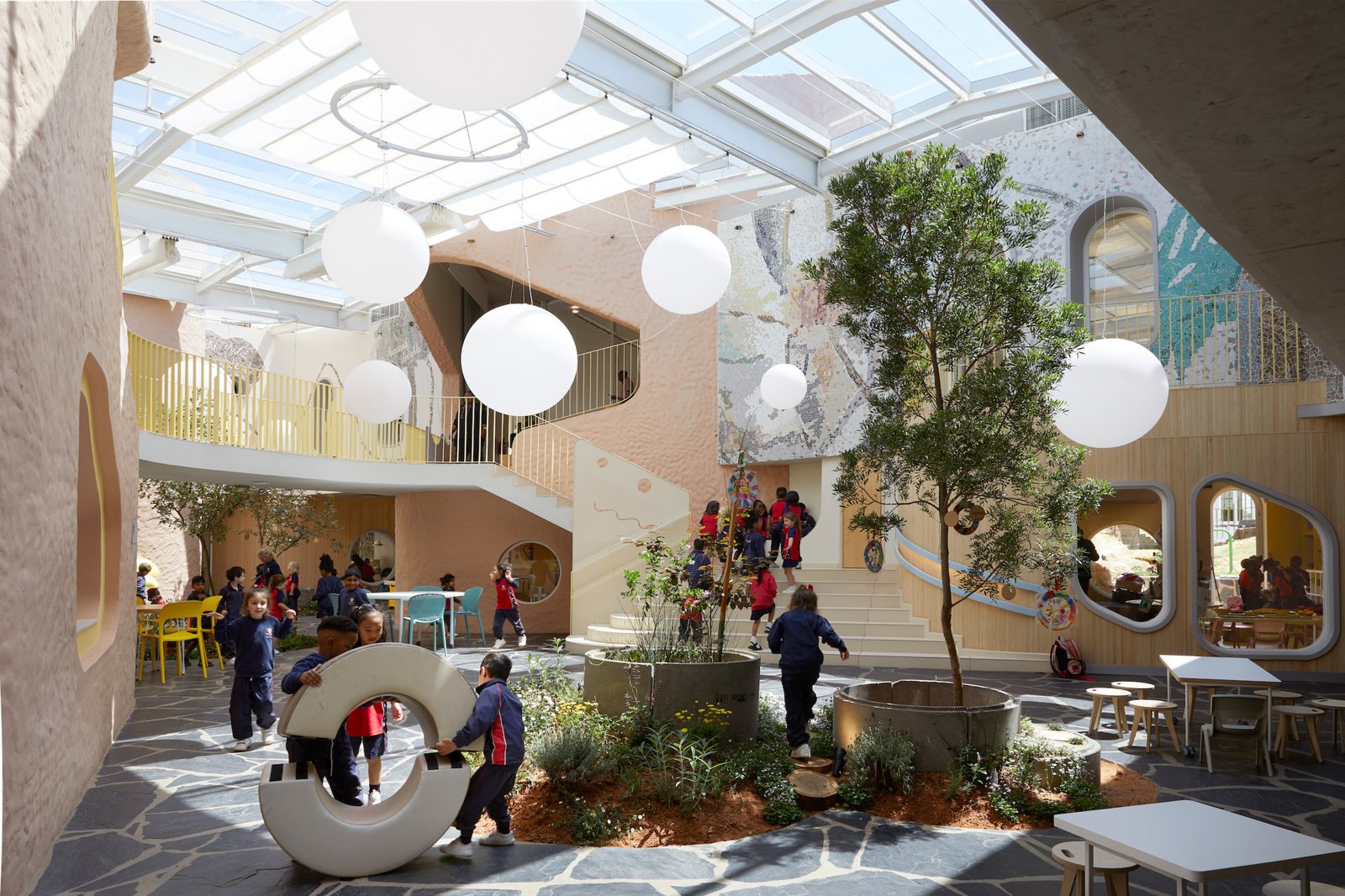 유리 지붕 아래 둥근 펜던트 조명이 떠 있는 Redhill Early Learning Centre 중앙 광장 The Piazza