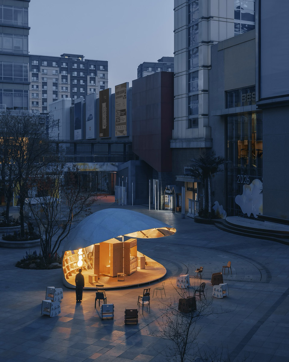 해 질 녘 상가 건물 사이에서 불을 밝힌 Shell Book Pavilion 외관