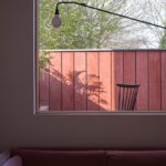 창 너머 레드 외장 벽과 나무가 보이는 Twin House 거실 창